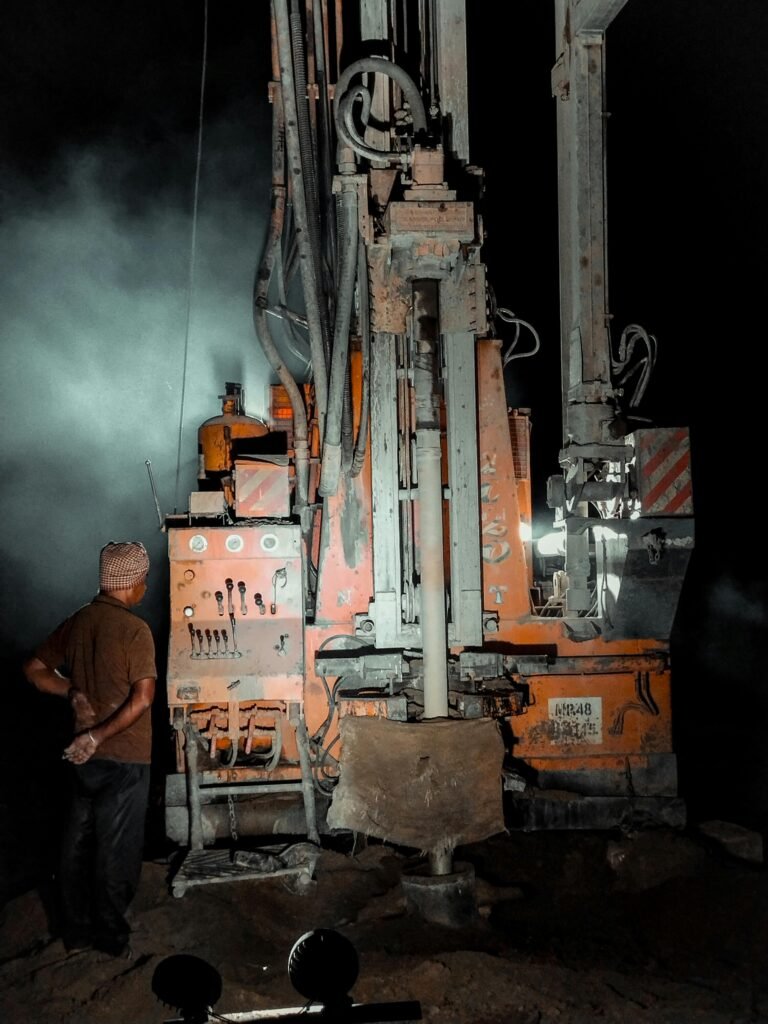 A worker operates a water well drilling rig under illumination during nighttime.