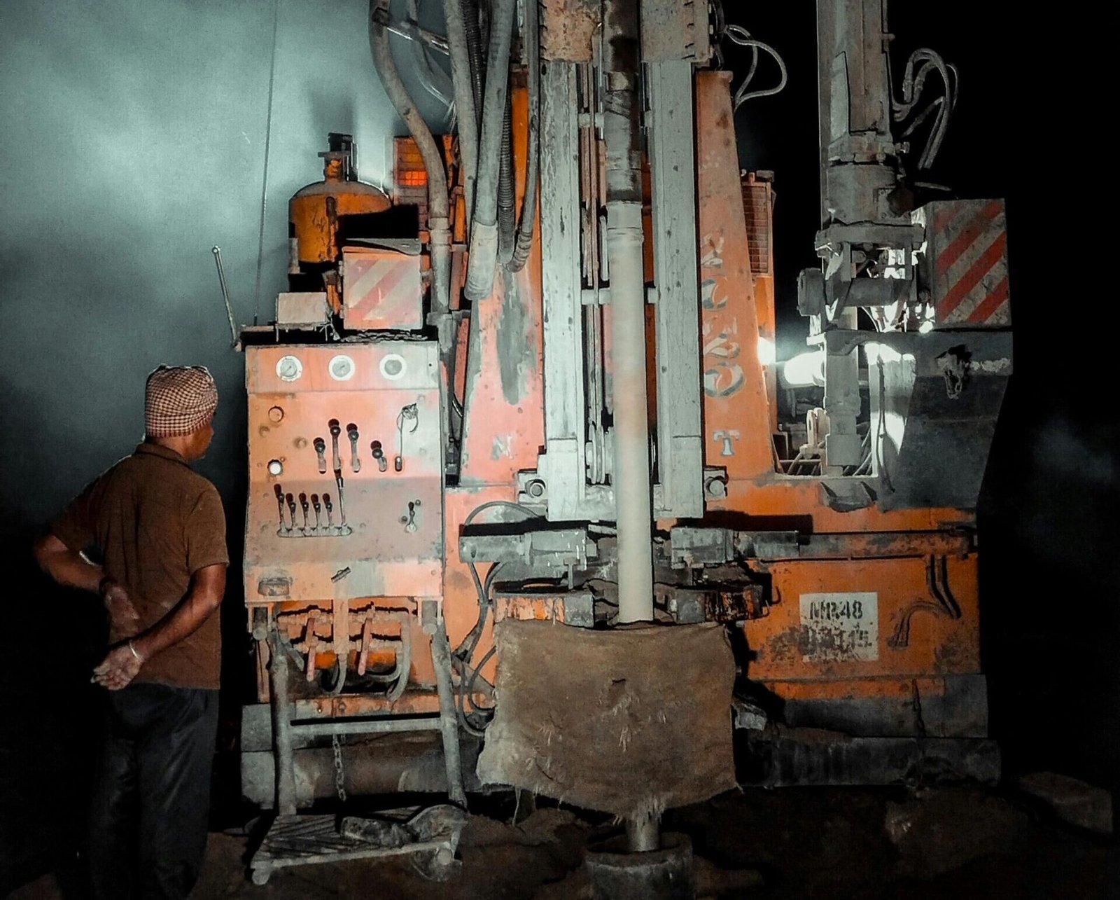 A worker operates a water well drilling rig under illumination during nighttime.